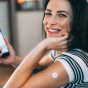 А woman smiling while holding her smartphone, with a sensor attached to her arm, likely for measuring blood sugar levels. She appears to be reviewing data or graphs from a health-related app on her phone. This scene captures the intersection of technology and personal health management, highlighting the importance of monitoring health metrics in a modern lifestyle. The woman's expression conveys positivity and engagement, making it an ideal representation of proactive health awareness.